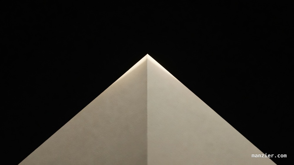 Extreme macro of a single sheet of cream paper folded into a sharp mountain fold under raking amber light against black